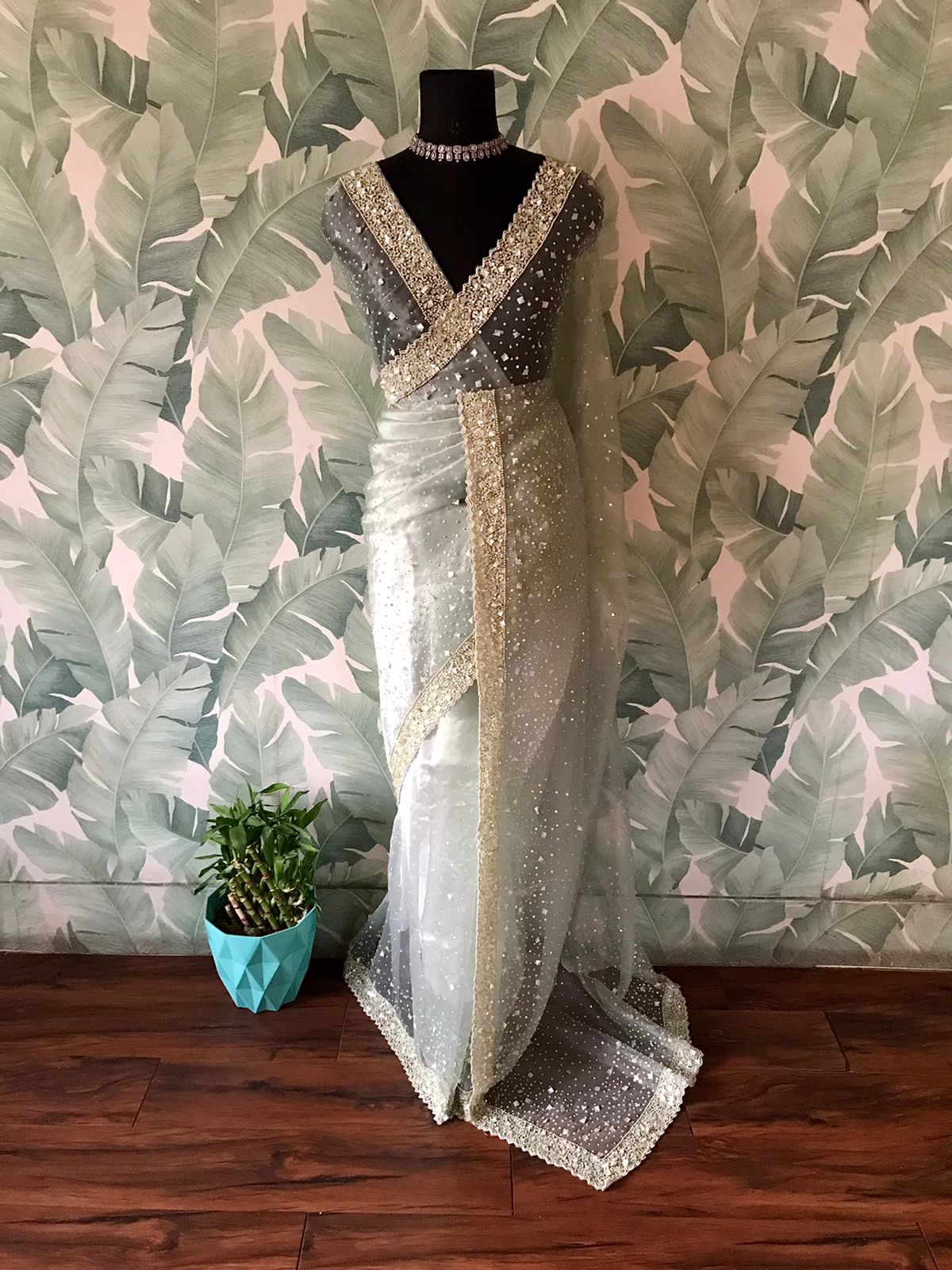 An elegant green organza-adorned tissue silk saree on a mannequin against a leaf-patterned wall.