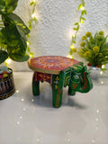 Decorative green and red elephant stool with a colorful design, painted with metallic paint on a white surface with greenery and lights in the background.